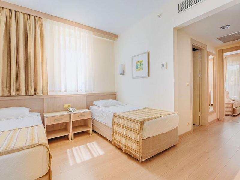 Bright hotel room with two single beds, wooden floor, and beige curtains.