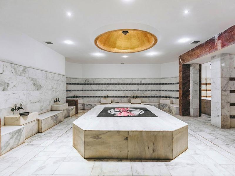 Spacious marble spa room with central massage table, ornate golden ceiling light, and wall shelves.
