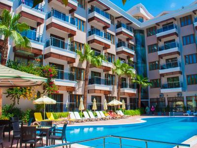 Hotel moderno con piscina all'aperto, ombrelloni, lettini e palme in una giornata soleggiata.
