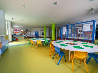 Bunter, heller Klassenraum mit runden Tischen, kleinen Stühlen und Spielzeugregalen an den Wänden.