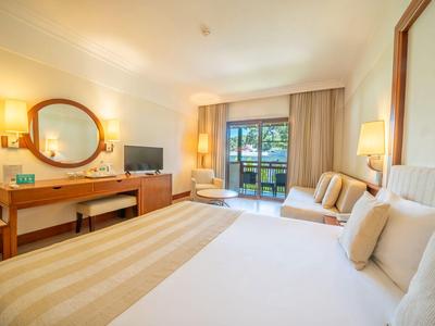 Chambre d'hôtel lumineuse avec grand lit, bureau, coin salon et balcon donnant sur l'eau.