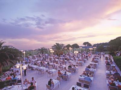 Große Terrasse mit vielen Tischen, Menschen sitzen zum Abendessen bei farbigem Sonnenuntergangshimmel.