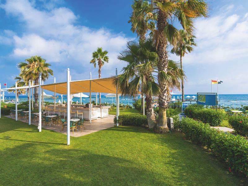 Jardín de hotel con césped, palmeras y zona de descanso con sombra junto al mar.