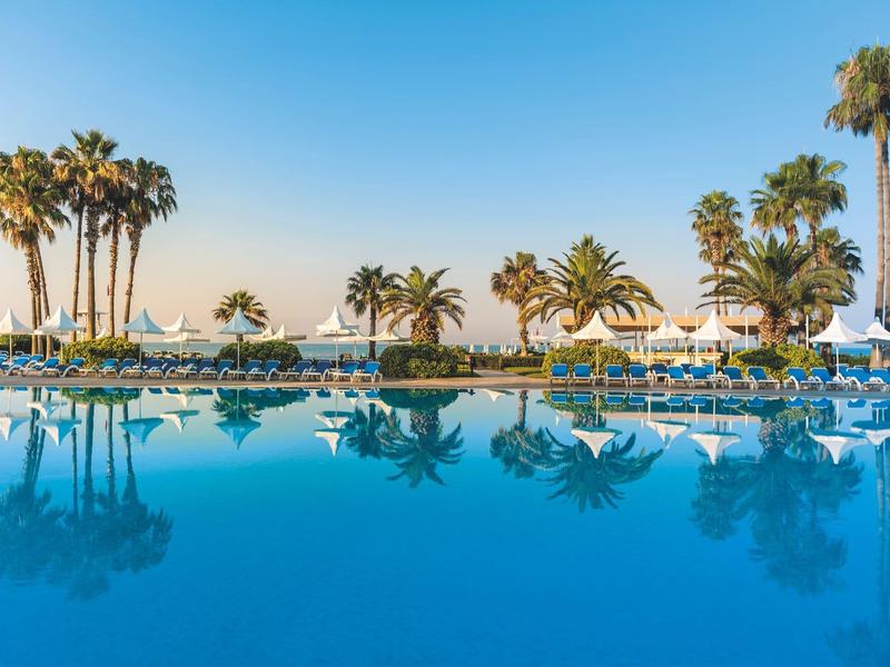 Una gran piscina de hotel con palmeras y tumbonas bajo un cielo azul claro junto al mar.