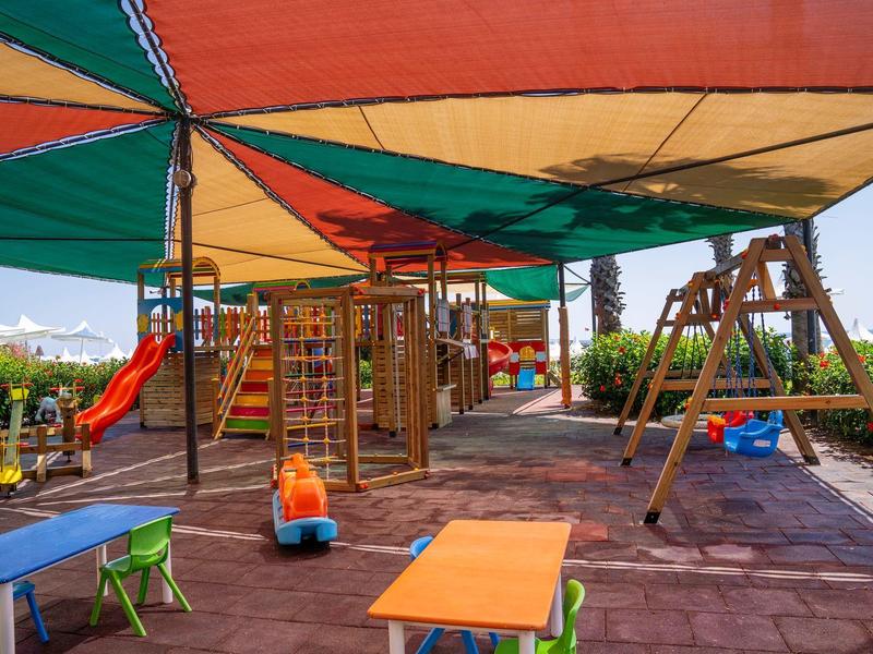 Área de juegos al aire libre con columpios, tobogán y mesas bajo toldos coloridos.