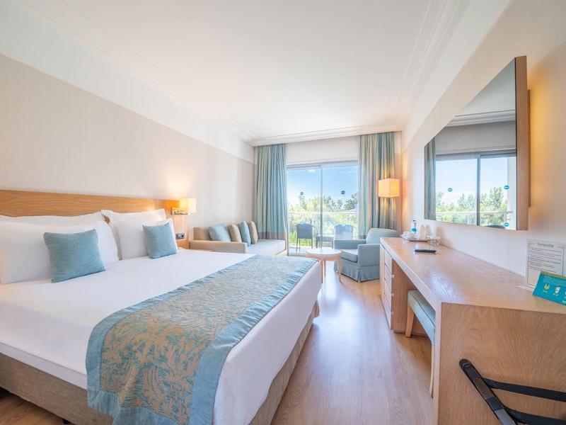 Habitación de hotel luminosa con cama grande, área de estar y vista al exterior a través de ventanas.