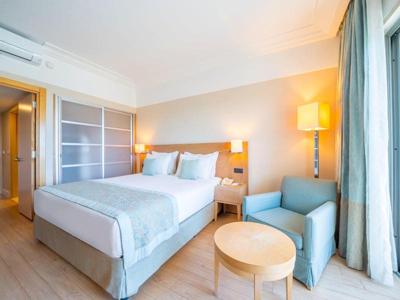 Habitación de hotel moderna con cama grande, área de descanso y luz natural por ventanas de piso a techo.