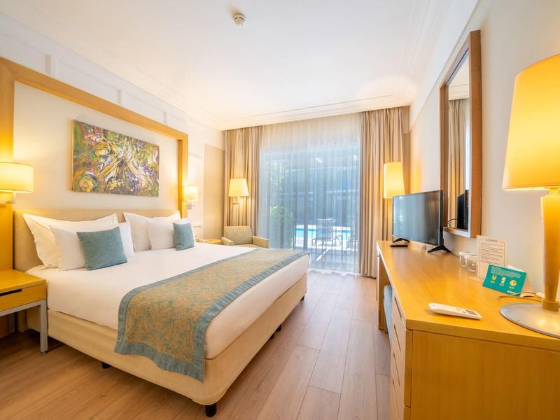 Habitación de hotel luminosa con cama grande, escritorio, TV y ventana panorámica con cortinas.