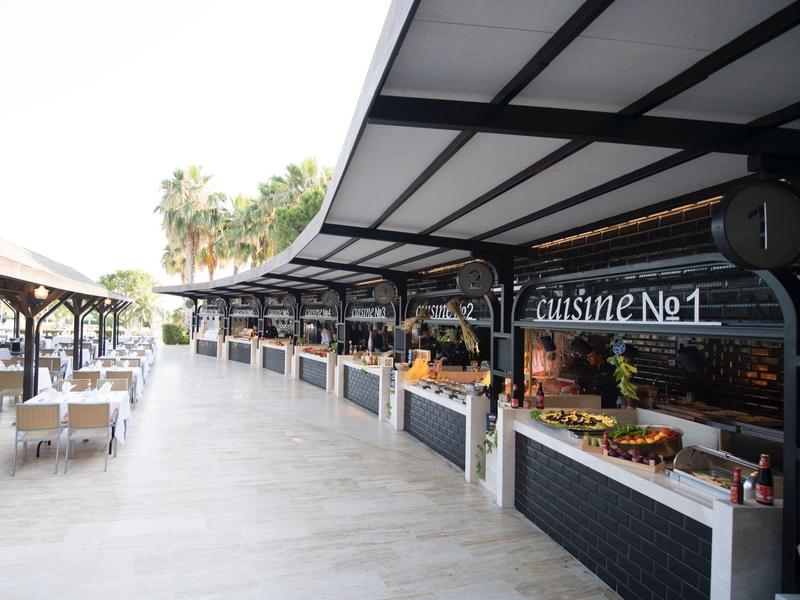 Área exterior moderna de un hotel con puestos cubiertos y asientos, palmeras al fondo.