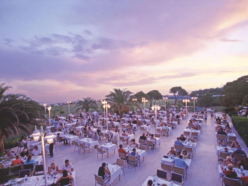 Terraza nocturna de hotel con muchos huéspedes y camino bordeado de palmeras al atardecer.
