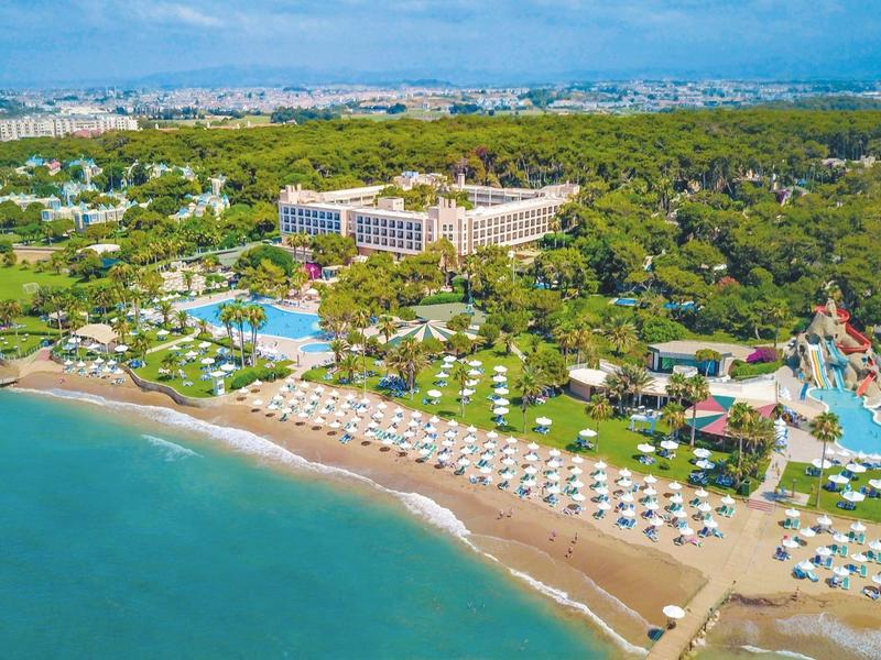 Gran hotel en la playa con tumbonas y sombrillas, rodeado de paisaje verde.
