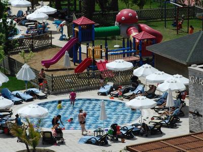 Buitenzwembad met mensen die zwemmen en zonnen, omgeven door ligstoelen en parasols bij een speeltuin.