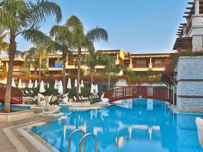 Hotelzwembad met ligstoelen, witte parasols en palmbomen onder een heldere lucht.