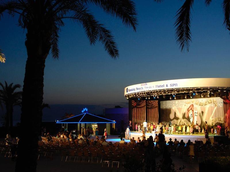 Openluchtconcert 's nachts met palmbomen en publiek aan het strand onder donkerblauwe lucht.
