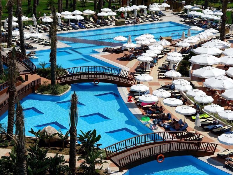 Groot hotelzwembad met meerdere bruggen, ligstoelen en witte parasols onder palmbomen.