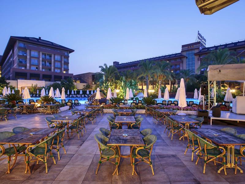 Buitenterras van hotel met veel tafels en parasols bij een zwembad in de schemering.