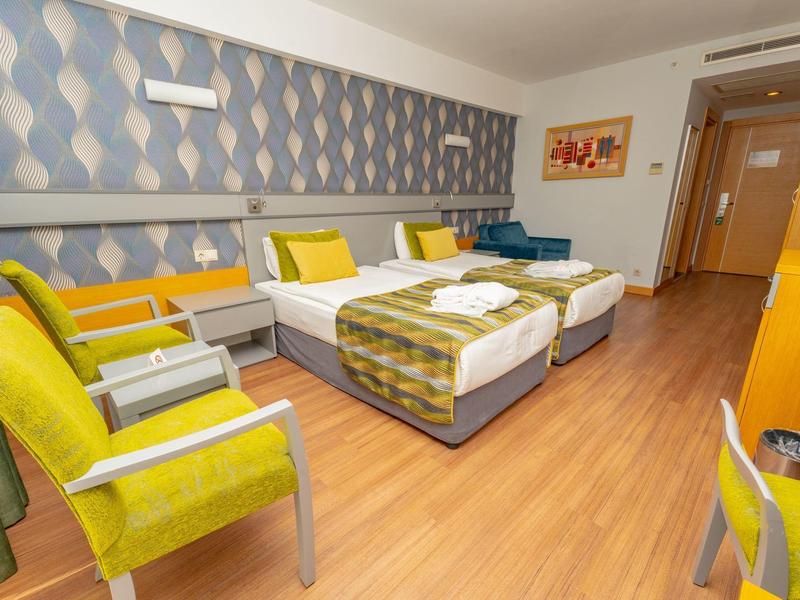 Licht hotelkamer met twee bedden, gele stoelen en houten vloer.