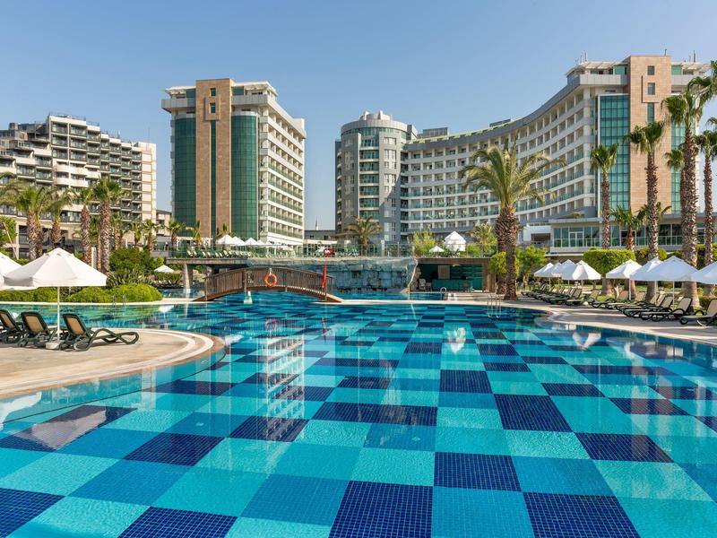 Basen w niebieską szachownicę z leżakami i nowoczesnym hotelem w tle.