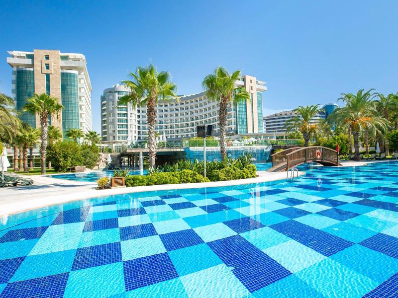 Basen w niebieskie kwadraty przed dużym hotelem otoczonym palmami.
