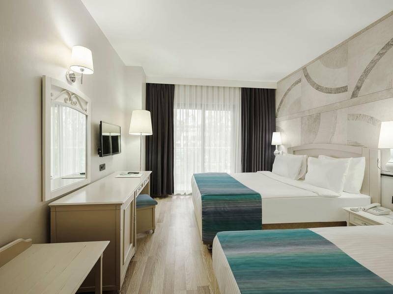 Habitación de hotel luminosa con dos camas, escritorio, TV y grandes ventanas con cortinas.