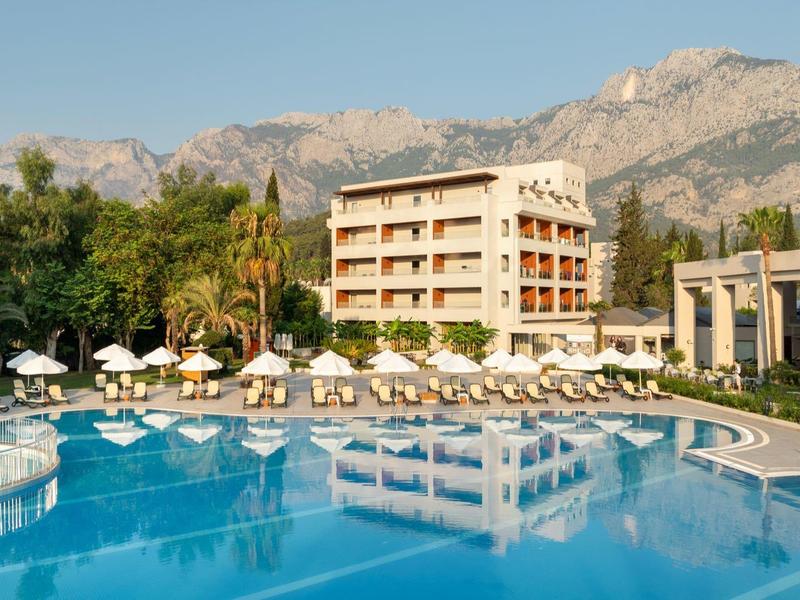 Hotel con gran piscina, sombrillas y montañas al fondo bajo cielo despejado.