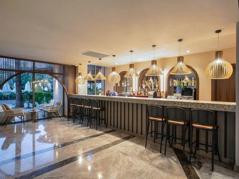 Área moderna de bar en hotel con taburetes elegantes y gran espejo detrás de la barra.