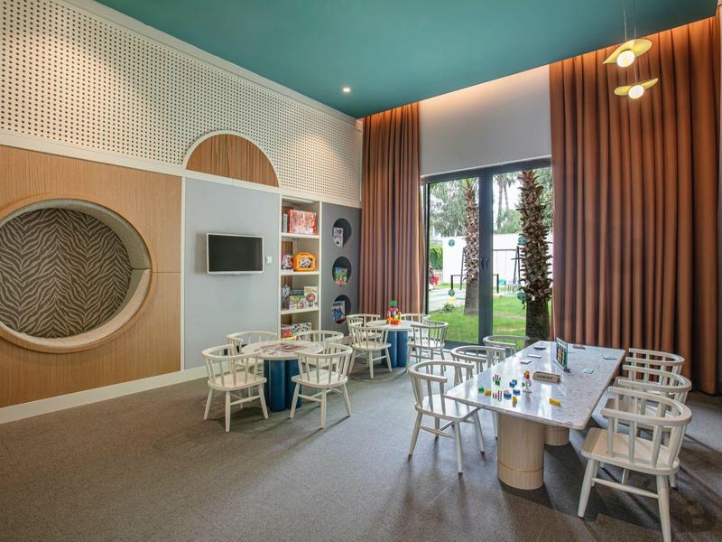 Área de salón moderna del hotel con grupos de asientos, grandes ventanas y cortinas con vistas al jardín.