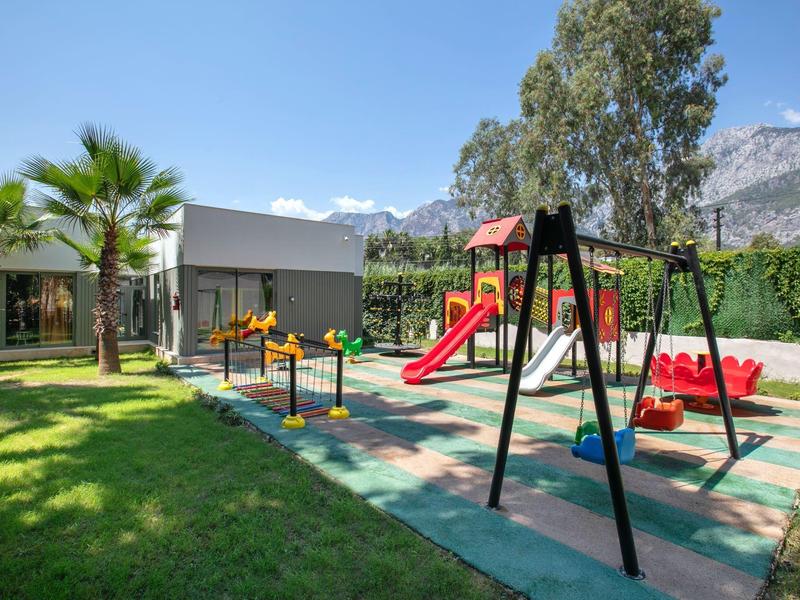 Área de juegos para niños al aire libre junto a un hotel, con columpios, toboganes y árboles alrededor.