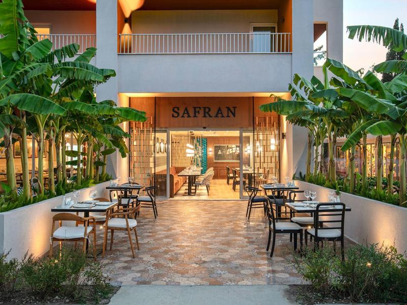 Área exterior moderna de un hotel con mesas, sillas y plantas verdes frondosas a ambos lados.