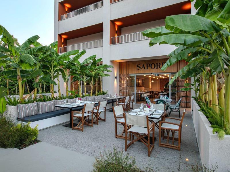 Área exterior moderna de un hotel con comedor, mesas de madera y plantas tropicales.