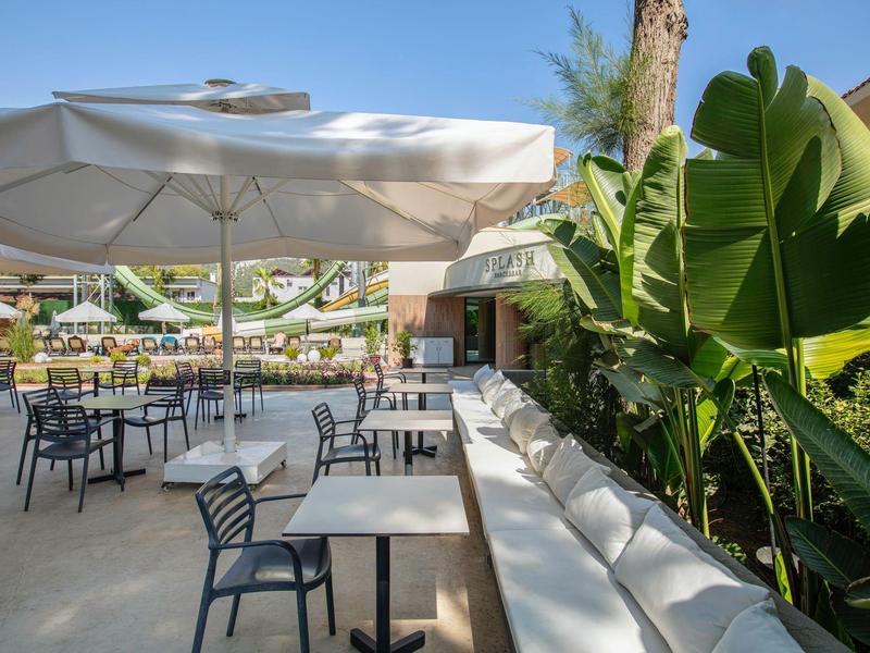 Terraza soleada con mesas, sillas y grandes sombrillas junto a plantas verdes.