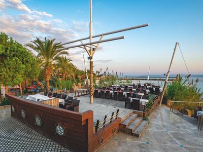 Restaurante al aire libre con palmeras y vista al mar, mesas y sillas en una terraza al atardecer.