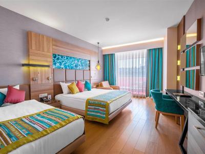 Modernes Hotelzimmer mit zwei Betten, bunten Kissen, Holzboden und türkisfarbenen Vorhängen.