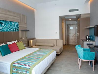 Habitación de hotel moderna con cama, sofá, escritorio y sillas en tonos neutros.