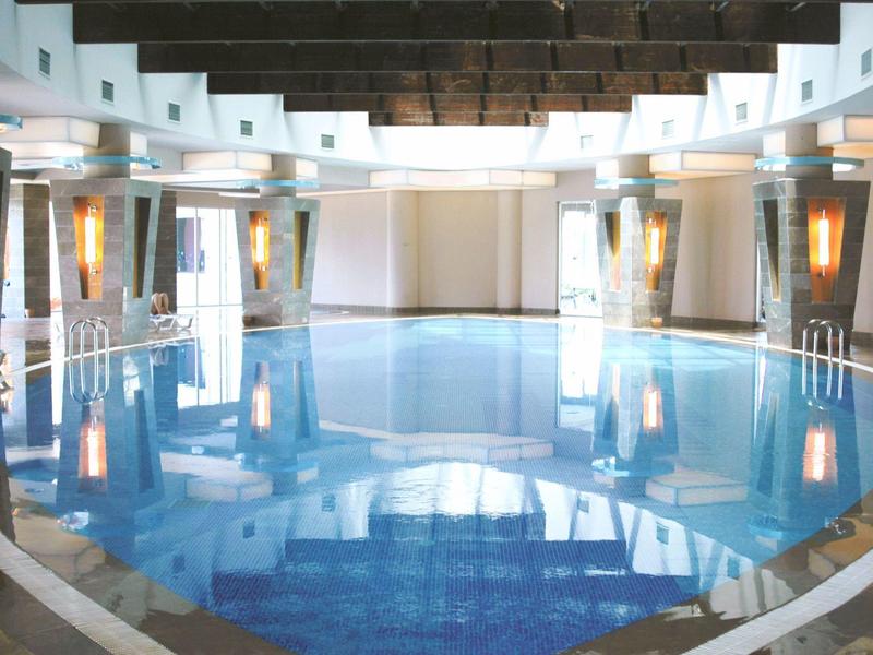 Piscina interior moderna en un hotel con paredes blancas, techo alto y cálidos acentos de luz.