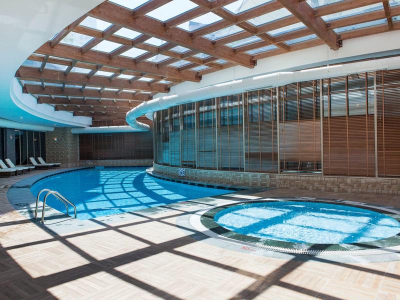 Área interior con piscina de hotel, cubierta de madera y jacuzzi adyacente con agua azul.