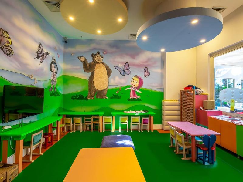 Sala de juegos infantil colorida con mesas, sillas y murales de personajes de dibujos animados