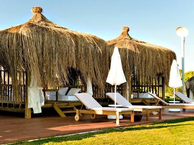 Twee ligstoelen met witte handdoeken onder rieten daken naast een groen gazon onder een blauwe lucht.