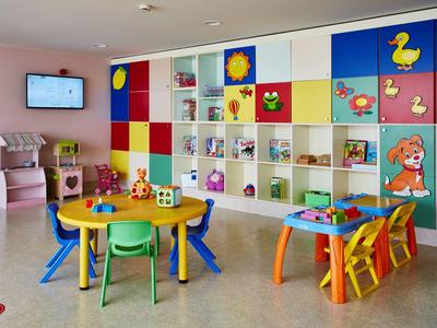 Kleurrijke speelkamer voor kinderen met tafels, stoelen, planken met speelgoed en felle muurdecoraties.