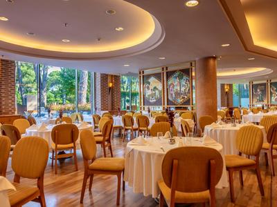 Elegantní restaurace s kulatými stoly, pohodlnými židlemi a velkými okny s výhledem na zeleň.