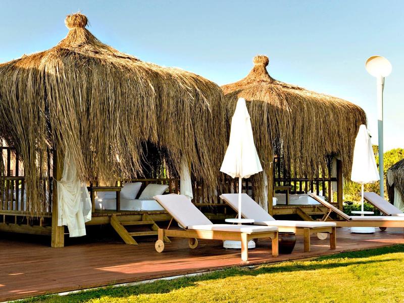 Twee ligstoelen met witte handdoeken onder rieten daken naast een groen gazon onder een blauwe lucht.