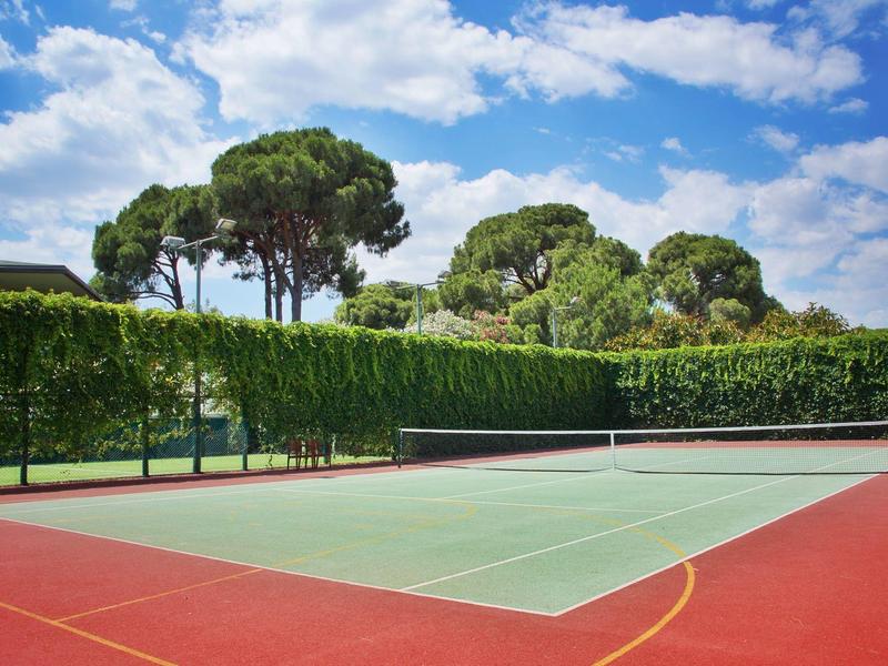 Buiten tennisbaan met groen oppervlak omringd door bomen en blauwe lucht met wolken.
