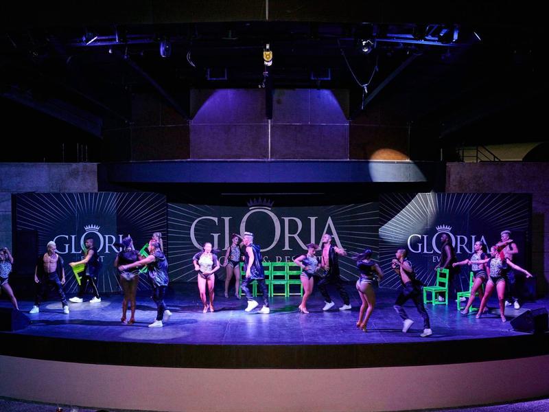 Podiumoptreden met dansers in kleurrijke kleding en groene stoelen bij Gloria-evenement.