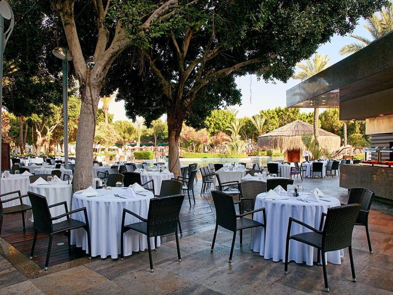 Przestrzeń restauracyjna na zewnątrz z stołami, białymi obrusami i czarnymi krzesłami pod drzewami.