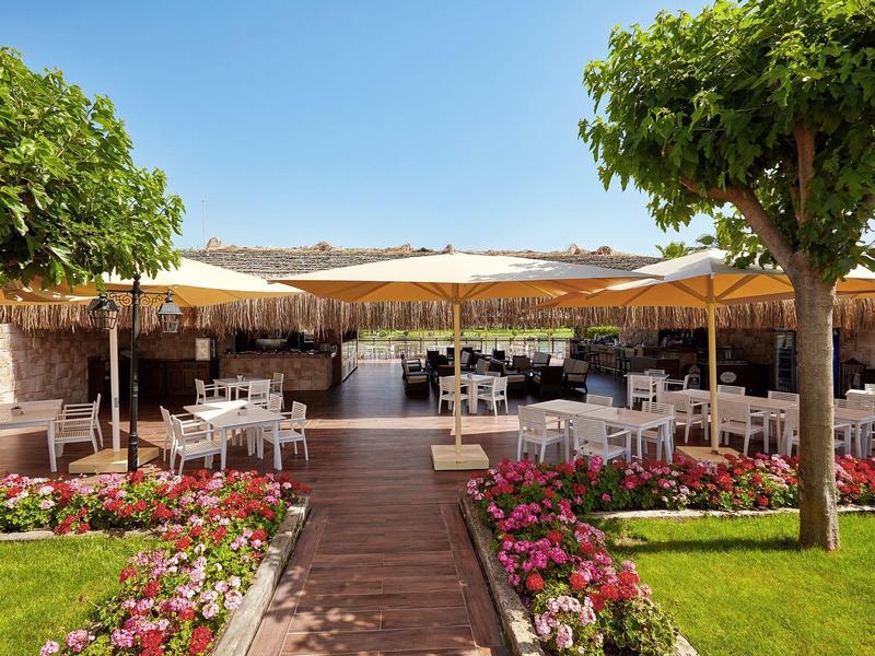 Taras hotelowy z białymi krzesłami, stołami i parasolami, otoczony kwiatami i drzewami.