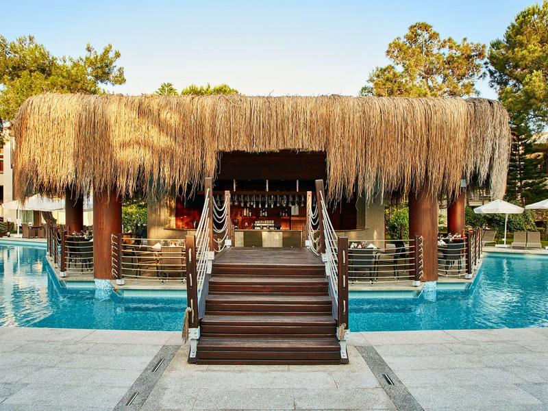 Bar przy basenie z dachem ze słomy, otoczony niebieskim basenem i zielonymi drzewami w tle.