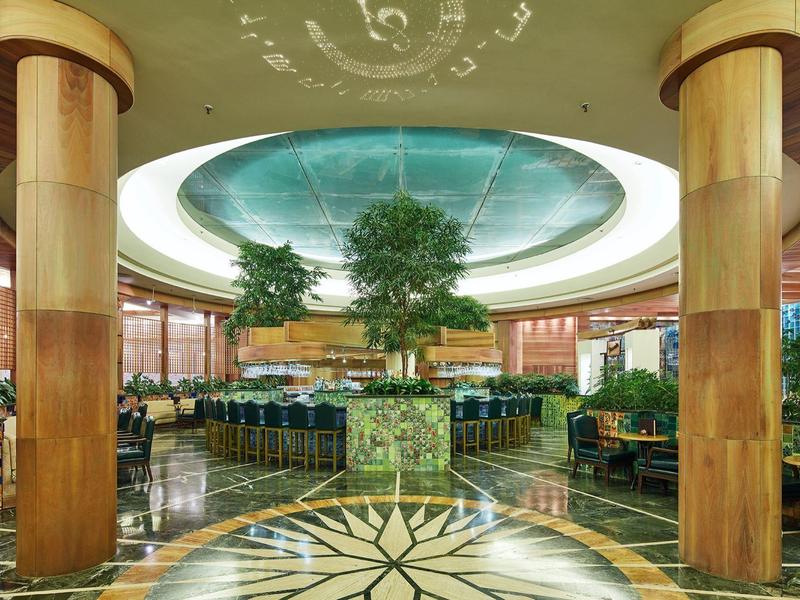 Przestronny hol hotelowy z marmurową podłogą, kolumnami i centralnymi drzewami pod oświetlonym sklepieniem kopuły.