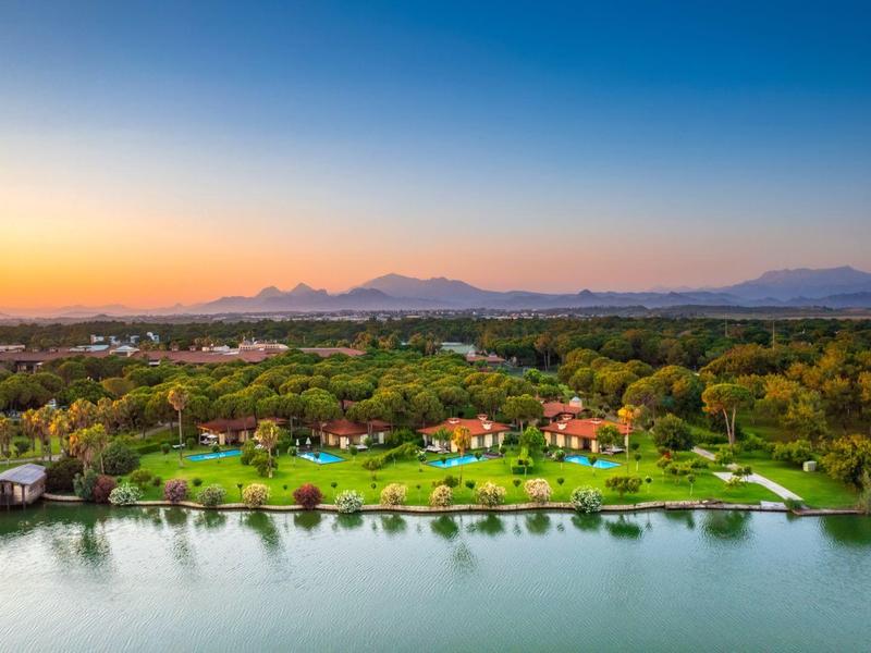 Krajobraz z jeziorem, zielonymi łąkami i budynkami o zachodzie słońca.