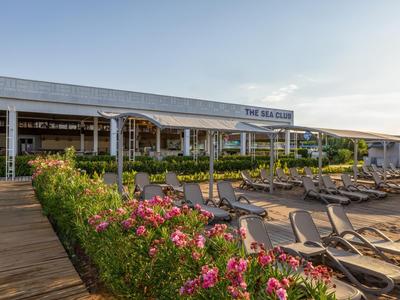 Wide wooden walkway leads to hotel with outdoor pool and lounge chairs under clear sky.