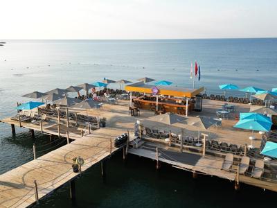 Large wooden pier with sunshades and seating areas over calm sea water under clear sky.